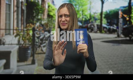 Femme tenant un passeport bleu, paume levée dans une rue urbaine animée avec des voitures et des vélos garés ; réticence. Banque D'Images