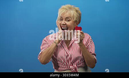 Femme hispanique senior tient un petit cœur rouge près de sa joue tout en riant et en souriant dans un studio bleu portant une chemise rayée rouge ; Joy love celea Banque D'Images