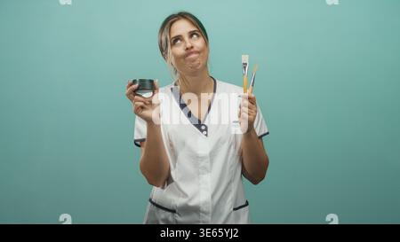 Femme tenant un pot de crème et des pinceaux, portant un uniforme blanc et levant les mains tout en levant les yeux en studio ; décision réfléchie. Banque D'Images
