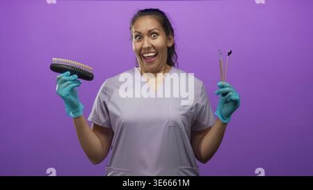 Femme dentiste souriant largement tenant guide de teinte de dent et miroir dentaire avec les mains gantées dans le studio violet ; confiance joyeuse. Banque D'Images