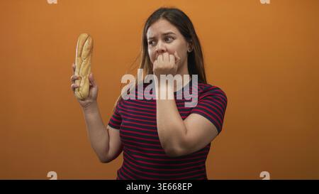 Femme tenant une baguette mord des ongles en studio avec fond orange et chemise rayée ; anxiété malaise hésitation. Banque D'Images