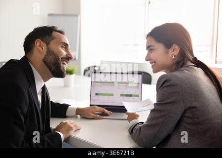 Membres de l'équipe multiethnique souriants assis à la table du bureau avec un ordinateur portable Banque D'Images