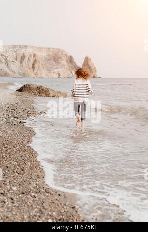 Femme qui court sur la plage femme insouciante en pull rayé court pieds nus à travers les vagues de l'océan peu profondes sur une belle côte rocheuse Banque D'Images