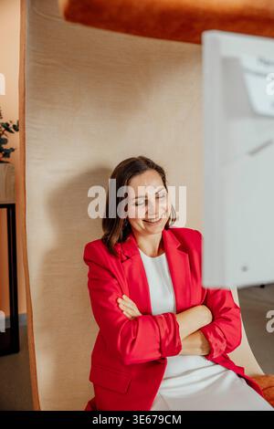 Rire jeune femme caucasienne avec les cheveux bruns est assis soigneusement dans l'espace de coworking moderne. Elle porte un blazer rouge et a une posture détendue Banque D'Images
