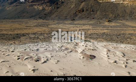 Vue aérienne d'une plage de sable rencontrant broussailles arides et falaises rocheuses avec un véhicule garé près des dunes, vaste paysage aride sous la lumière du jour. Banque D'Images