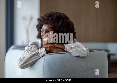 Femme d'affaires adulte moyenne positive avec coiffure afro regardant loin réfléchi tout en se relaxant sur le canapé dans le bureau Banque D'Images