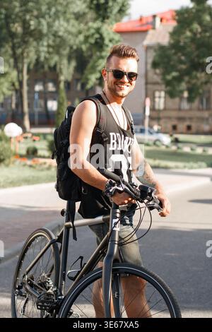 Heureux homme beau hipster avec des lunettes de soleil dans le débardeur se tient près d'un vélo et s'amuser dans la ville sur une journée ensoleillée Banque D'Images