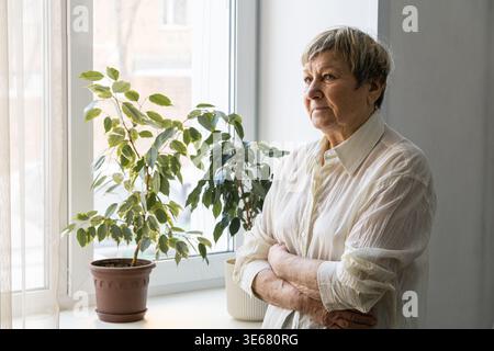 Femme senior debout, les bras croisés, regardant réfléchi à travers une fenêtre avec des plantes d'intérieur sur le seuil. Femme âgée contemplant la vie Banque D'Images