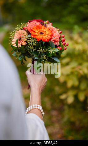 Mariée tenant un bouquet de mariage d'automne vibrant avec gerberas orange et roses. Gros plan des mains avec bracelet en perles sur fond de jardin flou Banque D'Images