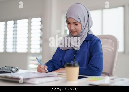 Femme musulmane asiatique diligente se concentrant sur la paperasse. L'apprenant professionnel effectue l'analyse des données en écrivant des notes sur les documents au bureau de travail. Banque D'Images