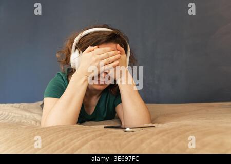 Femme portant des écouteurs et les mains couvrant ses yeux, couchée sur un lit se sentant submergée par l'anxiété et la vie moderne, faisant face au stress Banque D'Images