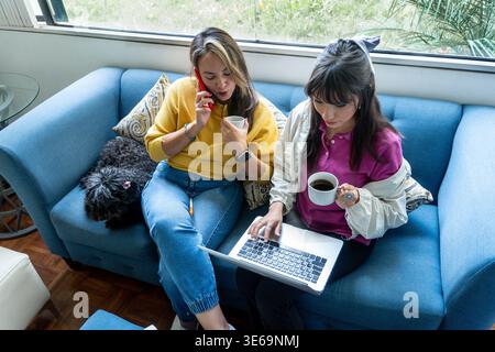 Deux femmes travaillant, utilisant un ordinateur portable, parlant au téléphone et buvant du café sur un canapé Banque D'Images