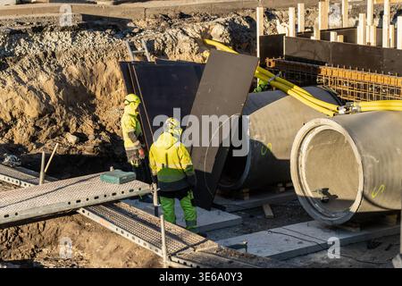 Chantier souterrain de construction de services publics avec des ouvriers installant des tuyaux d'égout en béton et des conduits jaunes de gaz ou d'électricité dans une tranchée profonde pour l'intérieur urbain Banque D'Images