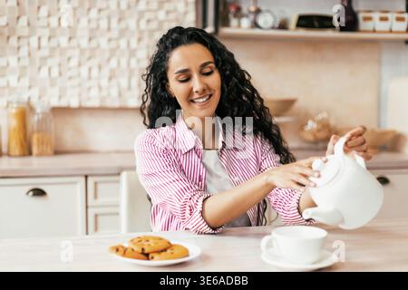 Femme verse du thé tout en étant assis à la table de cuisine avec des cookies sur une assiette pendant l'après-midi Banque D'Images