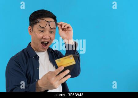 Homme asiatique surpris lève ses lunettes en regardant un smartphone et une carte de crédit sur fond bleu, idéal pour les alertes de paiement, les achats en ligne, Banque D'Images