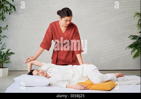 Masseuse faisant massage yoga thaïlandais. Thérapeute de femme faisant le traitement de massage traditionnel, étirant le corps de la patiente féminine. Client allongé sur tapis, profitant d'un massage des mains dans une atmosphère calme. Banque D'Images