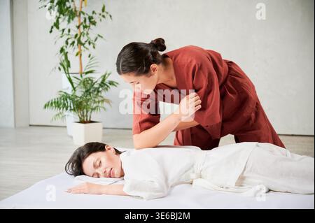 Masseuse faisant massage yoga thaïlandais. Thérapeute de femme faisant le traitement de massage traditionnel pour la patiente féminine utilisant le coude. Client allongé sur tapis, méditant, profitant d'un massage des mains dans une atmosphère calme. Banque D'Images