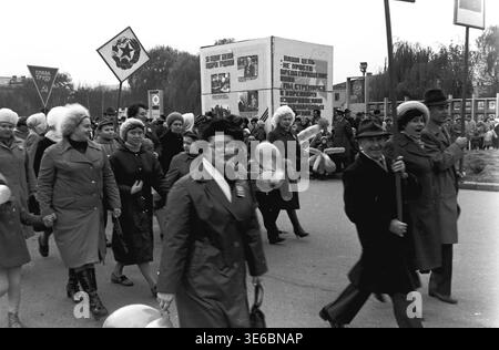 Slaviansk, 7 novembre 1983, place de la Révolution d'octobre (aujourd'hui Soborna). Une photo d'archive capturant un groupe de manifestants souriants pendant le défilé. Les femmes en manteaux et chapeaux chauds tiennent de longs ballons et marchent joyeusement. Un grand cube informatif avec des textes politiques et des portraits se tient derrière eux sur la place. Un signe carré avec une étoile soviétique, un marteau et une faucille est maintenu sur un poteau. La zone est remplie de marcheurs et d'arbres par temps d'automne. C'est une scène de vie paisible dans le Donbass. Banque D'Images