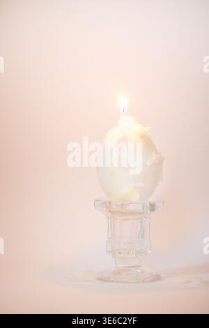 Bougie blanche en forme d'oeuf avec une flamme allumée se trouve sur un support en verre transparent, entourée d'un fond doux et neutre, soulignant la simplicité et l'élégance Banque D'Images