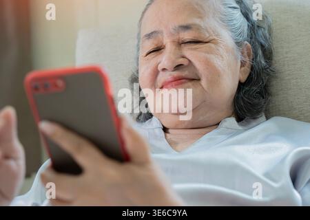 Gros plan d'une femme asiatique senior utilisant un smartphone sur un canapé à la maison. Mise au point sélective. Communication, internet et parler sur smartphone concept. Banque D'Images