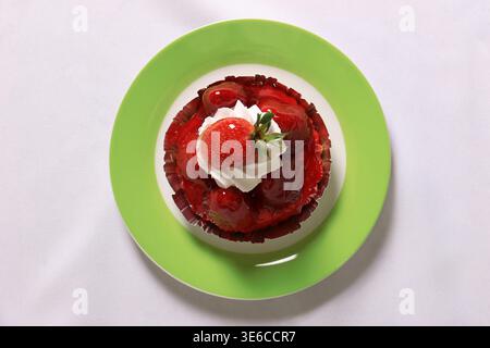 Petite tarte ronde aux fraises garnie de crème fouettée servie sur une assiette verte et blanche sur une nappe blanche. Banque D'Images