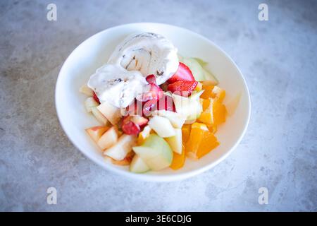 Assortiment de fruits en dés, dont melon, fraises et tranches d'orange, servi avec une boule de glace à la vanille dans un bol blanc sur une surface légère Banque D'Images