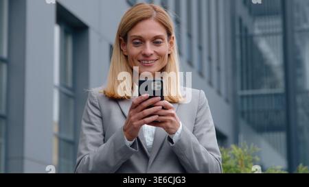 Heureuse femme d'affaires adulte caucasienne avec le téléphone appréciant le contenu en ligne dans la ville femme d'affaires d'âge moyen discutant le message électronique exécutif féminin Banque D'Images