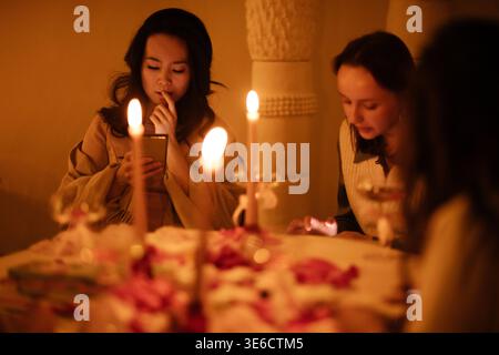 Deux femmes assises à une table éclairée aux chandelles avec des décorations florales, l'une regardant un smartphone tandis que l'autre examine la disposition de la table Banque D'Images