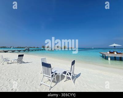 La scène montre du sable blanc, de l'eau claire et des villas sur l'eau aux Maldives. Des chaises et une table sont assis sur la plage, accueillant les visiteurs. Banque D'Images