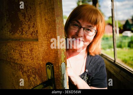 Femme d'âge moyen souriante regardant derrière une vieille porte en bois avec de la peinture fissurée dans la lumière chaude du soleil. Banque D'Images