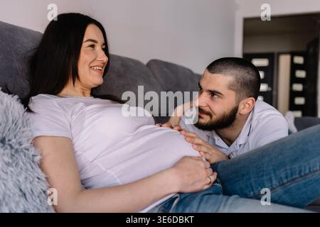 Portrait d'une femme enceinte heureuse tenant des chaussures de bébé et de son mari sur un canapé à la maison Banque D'Images