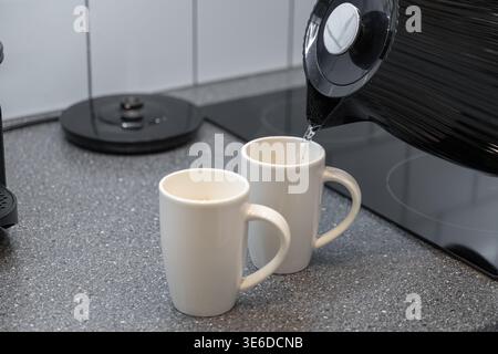 Gros plan d'une bouilloire électrique noire versant de l'eau vapeur dans une tasse en céramique blanche sur une cuisine moderne, mettant l'accent sur le confort quotidien et une boisson chaude Banque D'Images