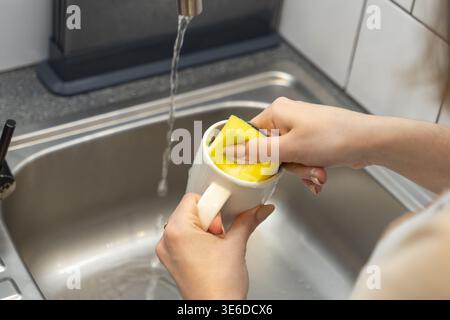 Mains frottant une tasse de café blanc avec une éponge jaune sous l'eau qui coule du robinet dans un évier de cuisine, illustrant le nettoyage domestique quotidien et hygi Banque D'Images