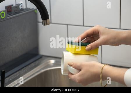 Vue détaillée des mains féminines lavant activement une tasse en céramique blanche avec une éponge jaune au-dessus d'un évier de cuisine, soulignant le nettoyage ménager essentiel Banque D'Images