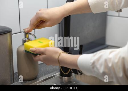 Un cliché détaillé capture les mains pressant un distributeur de savon, mettant du savon à vaisselle liquide vert sur une éponge de cuisine jaune pour un usage de nettoyage de routine Banque D'Images