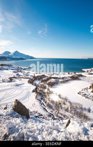 Vue aérienne d'un petit village côtier sur l'île de Kvaloya, en Norvège, entouré de montagnes enneigées et d'un fjord arctique calme sous un ciel hivernal clair Banque D'Images