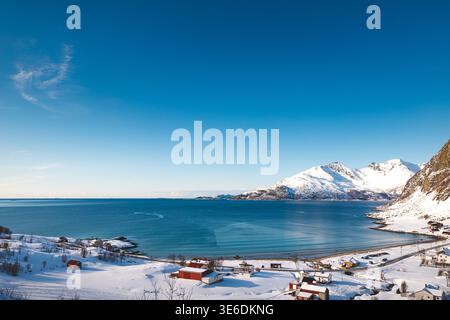 Vue aérienne d'un petit village côtier sur l'île de Kvaloya, en Norvège, entouré de montagnes enneigées et d'un fjord arctique calme sous un ciel hivernal clair Banque D'Images