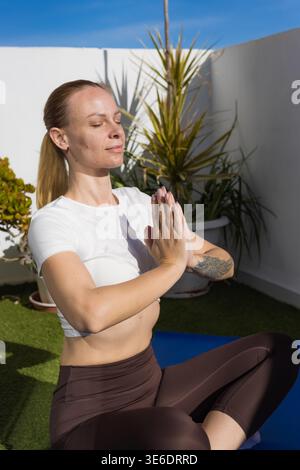 Femme assise les jambes croisées dans une pose méditative de yoga sur un tapis bleu à l'extérieur, pressant les mains ensemble au niveau de la poitrine avec les yeux fermés, embrassant le mindfu Banque D'Images