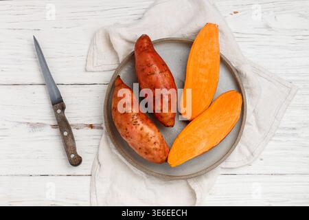 Patates douces entières et tranchées biologiques crues dans une assiette grise sur fond de bois blanc. Légumes racines frais. Mise au point sélective, vue de dessus et copie sp Banque D'Images