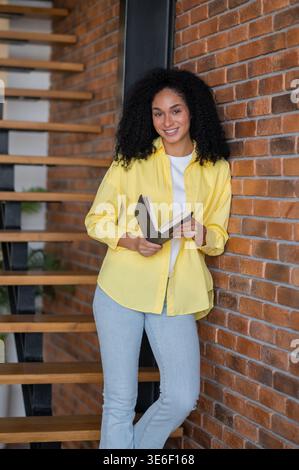 Mignonne femme souriante aux cheveux longs en veste jaune avec un livre dans les mains Banque D'Images