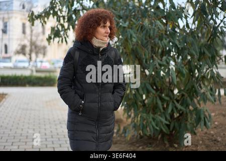 Femme mature avec les cheveux roux bouclés dans un manteau-gonflant foncé debout à l'extérieur dans un parc de la ville à côté d'un saule un jour d'hiver couvert Banque D'Images
