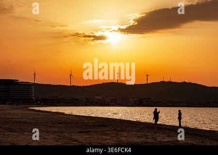 Coucher de soleil doré sur une baie balnéaire calme avec des silhouettes de gens sur la plage, des collines lointaines et des éoliennes à l'horizon, et des nuages oranges chauds Banque D'Images