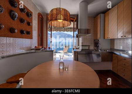 Cuisine élégante dans une luxueuse villa italienne au bord du lac, avec armoires en bois chaud, suspension tissée et portes ouvertes donnant sur une terrasse avec lac et moun Banque D'Images