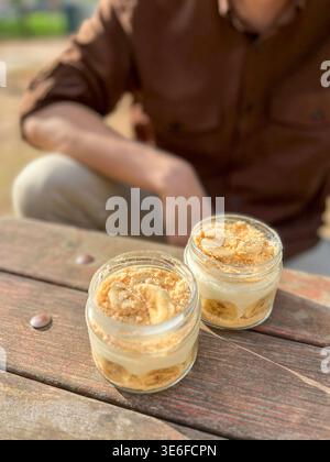Deux petits pots en verre remplis de pudding crémeux à la banane et de miettes de biscuits sont assis sur une table en bois rustique, stylisée dans une lumière naturelle chaude pour une maison confortable Banque D'Images