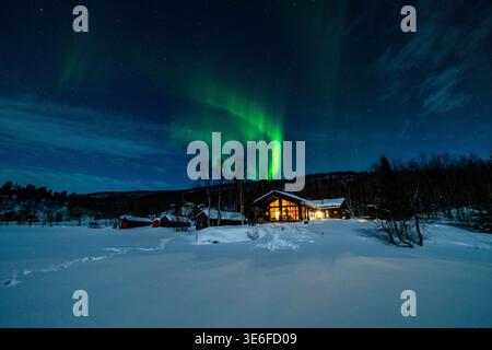 Green aurora borealis danse sur une cabane enneigée et un paysage forestier tranquille près de Tromso, en Norvège, en Laponie, sous un ciel étoilé clair. Banque D'Images