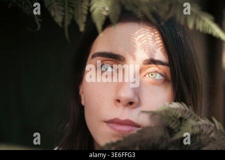Portrait en lumière naturelle d'une jeune femme entourée de feuilles de fougères avec ombre de lumière du soleil à motifs sur son front. Image féminine douce avec expres calmes Banque D'Images
