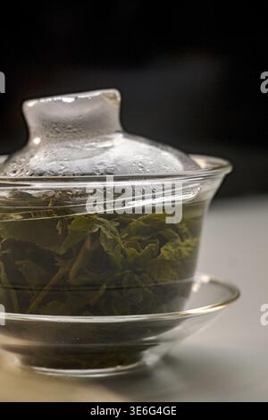 Il s'agit d'un gros plan de feuilles de thé fraîchement infusées placées dans une théière en verre transparent avec la vapeur qui monte légèrement Banque D'Images