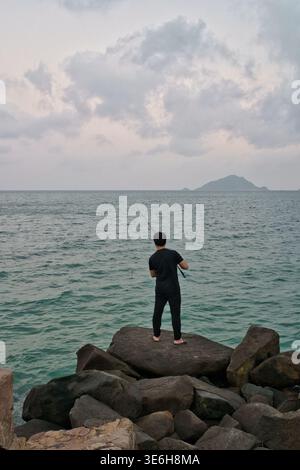 Un pêcheur solitaire se tient sur de grands rochers au bord de la mer, jetant sa ligne dans l'eau turquoise sous un ciel nuageux Banque D'Images