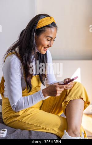 Femme asiatique adulte assise sur un canapé dans une salopette jaune, tenant un téléphone rose près d'un appareil blanc, souriant Banque D'Images