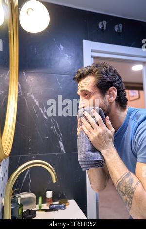 Homme se toilettant devant un miroir, essuyant son visage avec une serviette après le lavage, en se concentrant sur l'hygiène personnelle quotidienne Banque D'Images